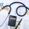 Image of a stethoscope, pen and calculator sitting atop charts and graphs