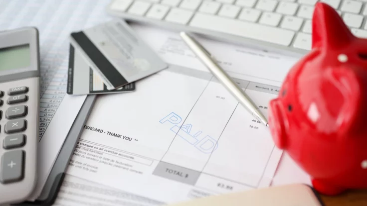 Image of a calculator, credit cards, pen and piggy bank sitting atop an invoice marked "Paid."