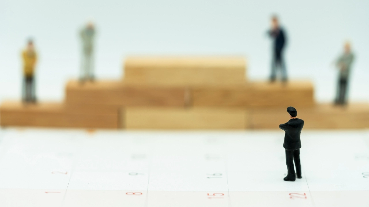 Image of miniature people standing on a wooden platform as a businessman assesses which one should be in top spot.