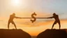 Image of two people, atop a mountain at sunset, pulling a rope with a dollar sign in the middle.