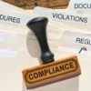 Image of a rubber stamp bearing the word "Compliance" sitting atop legal folders