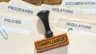 Image of a rubber stamp bearing the word "Compliance" sitting atop legal folders