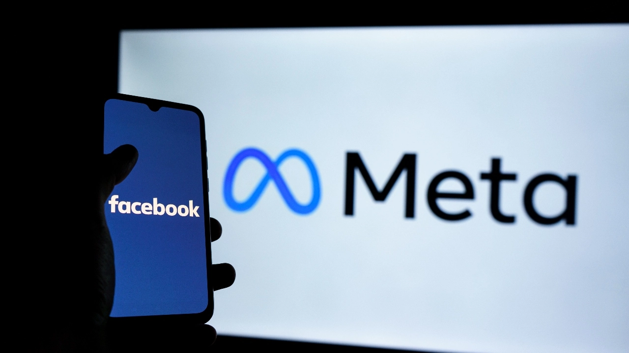 Image of a hand holding a cellphone bearing the Facebook logo in front of the Meta logo on a white screen